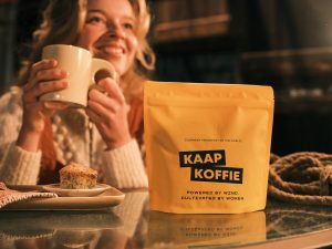 Smiling woman enjoying a cup of KAAP Koffie with a muffin, next to the yellow coffee bag. Organic sourced specialty coffee from Colombia.